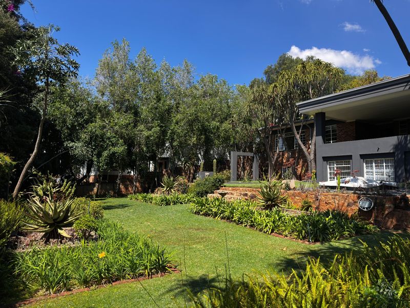 A well-maintained rear garden featuring a lush lawn, mature trees, stone retaining walls, and a covered patio area attached to the main house.