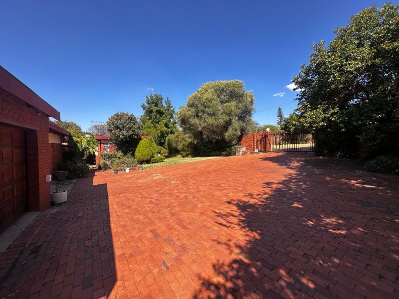 A spacious front exterior view featuring a large brick-paved driveway, secure metal entrance gate, and brick boundary walls with established garden landscaping.
