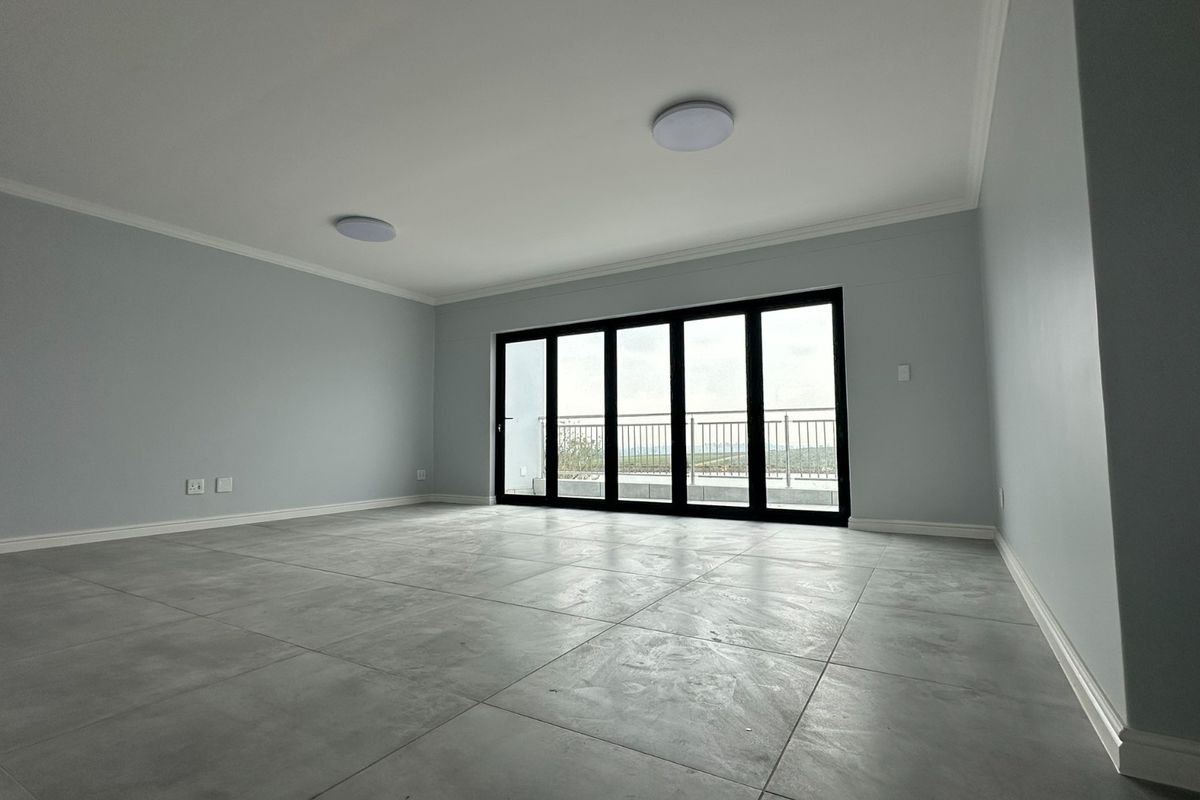 A spacious, empty living area featuring large format grey floor tiles, neutral walls, and black aluminium sliding doors leading to an outdoor balcony.