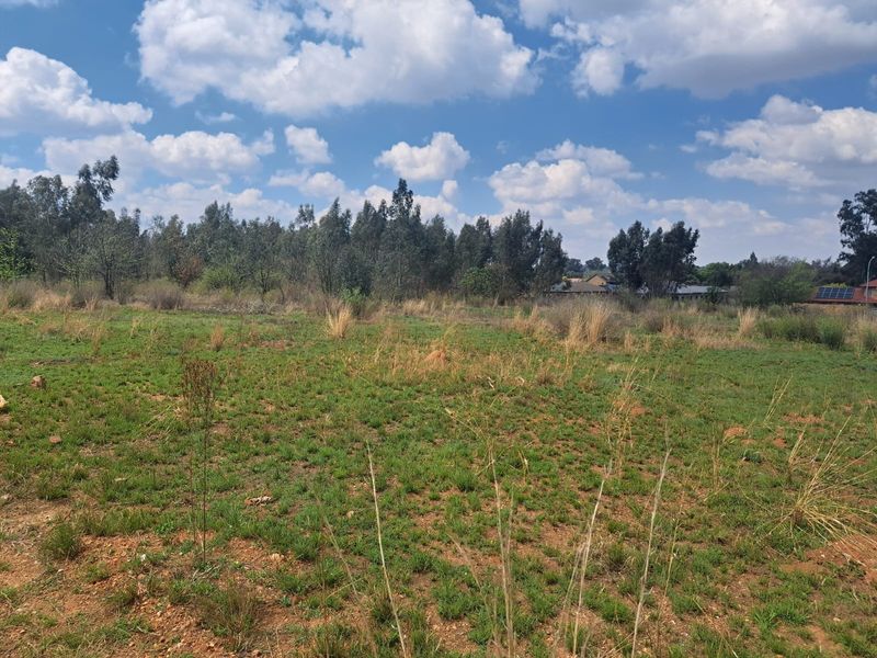 A view of a vacant residential plot featuring natural grass and surrounding trees under a clear blue sky.