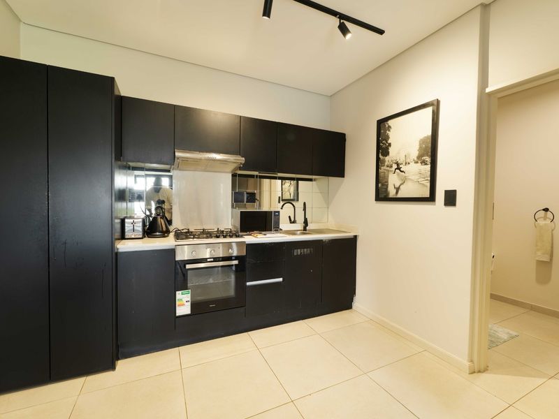 Modern kitchen featuring sleek black cabinetry, integrated gas hob, electric oven, extractor fan, and a mirrored splashback, complemented by neutral tiled flooring.
