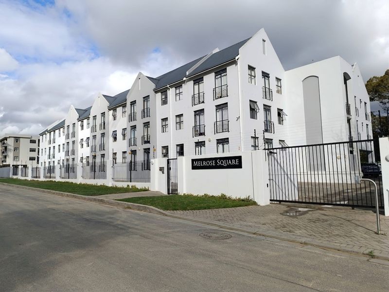 Street view of Melrose Square apartment complex featuring white facade, pitched roof, secure gated entrance, and perimeter electric fencing.