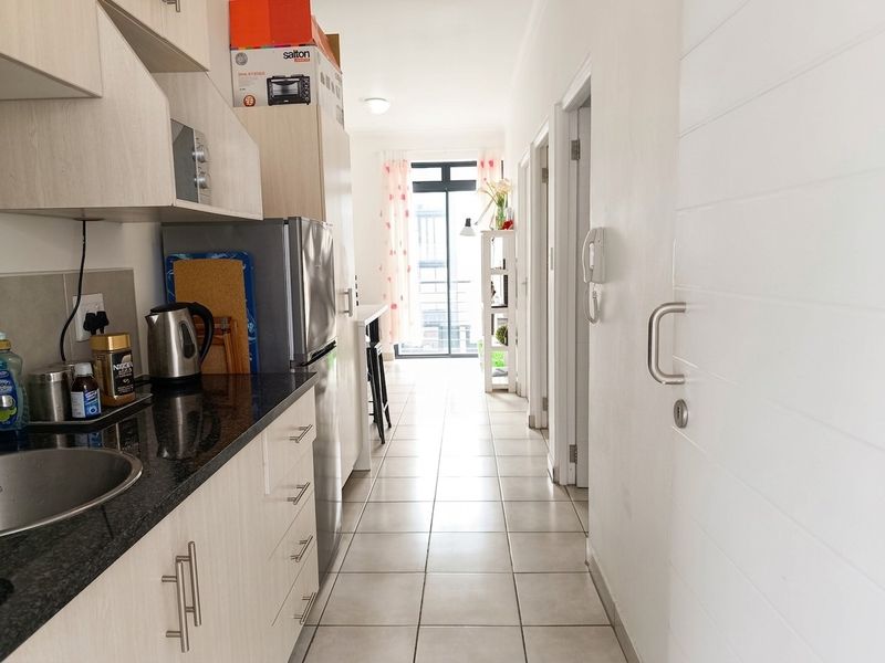 A compact kitchen featuring granite countertops, light wood-finish cabinetry, and tiled flooring, leading into a hallway with access to other rooms.