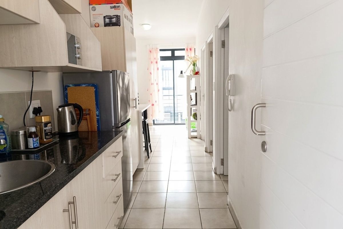 A compact kitchen featuring granite countertops, light wood-finish cabinetry, and tiled flooring, leading into a hallway with access to other rooms.