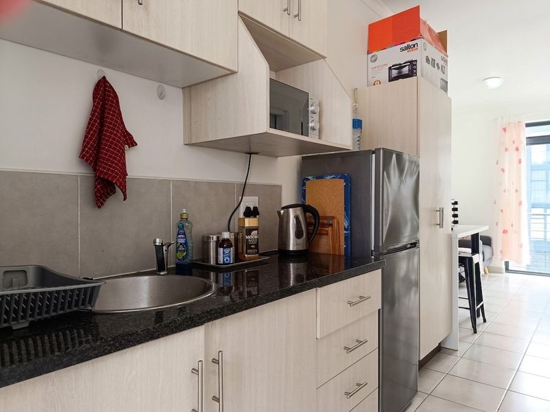 Compact kitchen featuring light wood-finish cabinetry, dark granite countertops, stainless steel sink, and tiled flooring, leading into an open-plan living area.