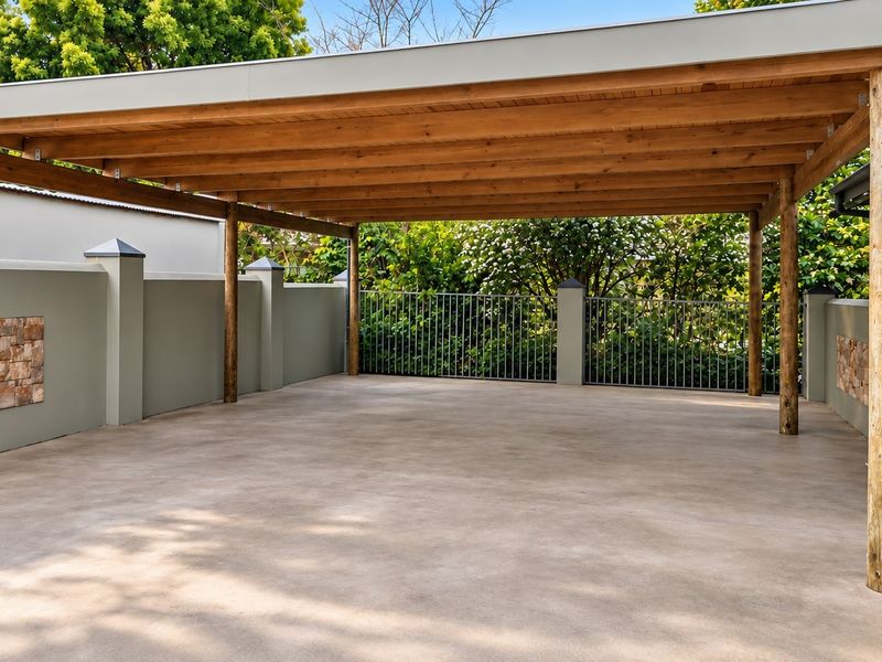 A well-maintained, open-sided carport featuring a timber roof structure, concrete flooring, and decorative stone-clad boundary walls.