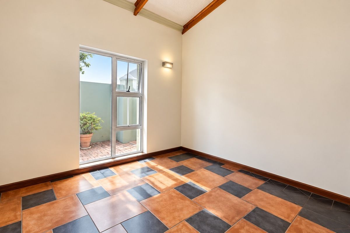 An empty living area featuring a vaulted ceiling with exposed timber beams, tiled flooring, and a large window providing natural light.