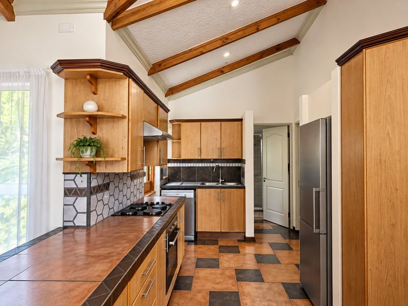 A well-maintained kitchen featuring wooden cabinetry, tiled countertops, built-in appliances, and distinctive exposed timber ceiling beams with tiled flooring.