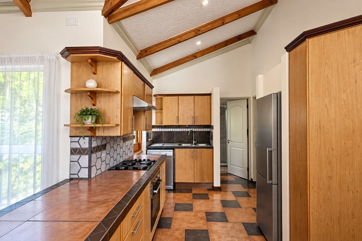 A well-maintained kitchen featuring wooden cabinetry, tiled countertops, built-in appliances, and distinctive exposed timber ceiling beams with tiled flooring.