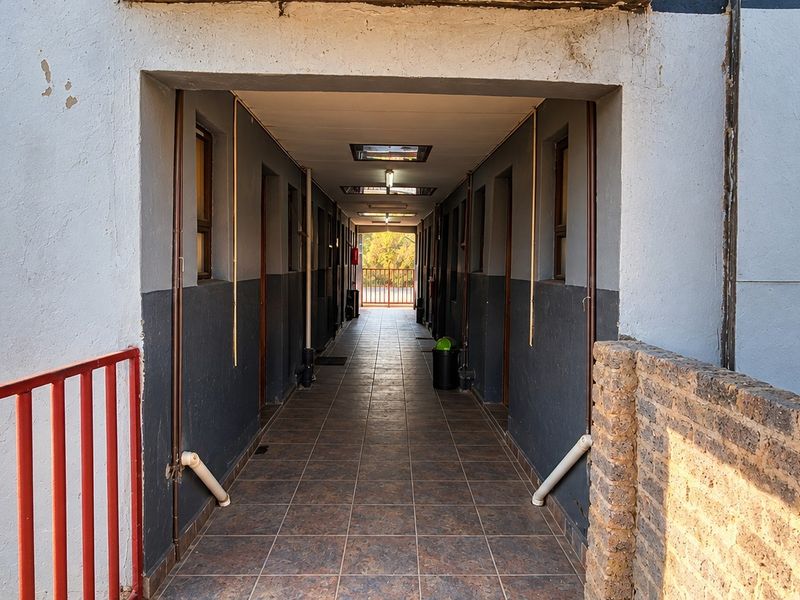 A long, tiled commercial hallway featuring recessed lighting, metal railings, and access to multiple units, showing signs of exterior wear.
