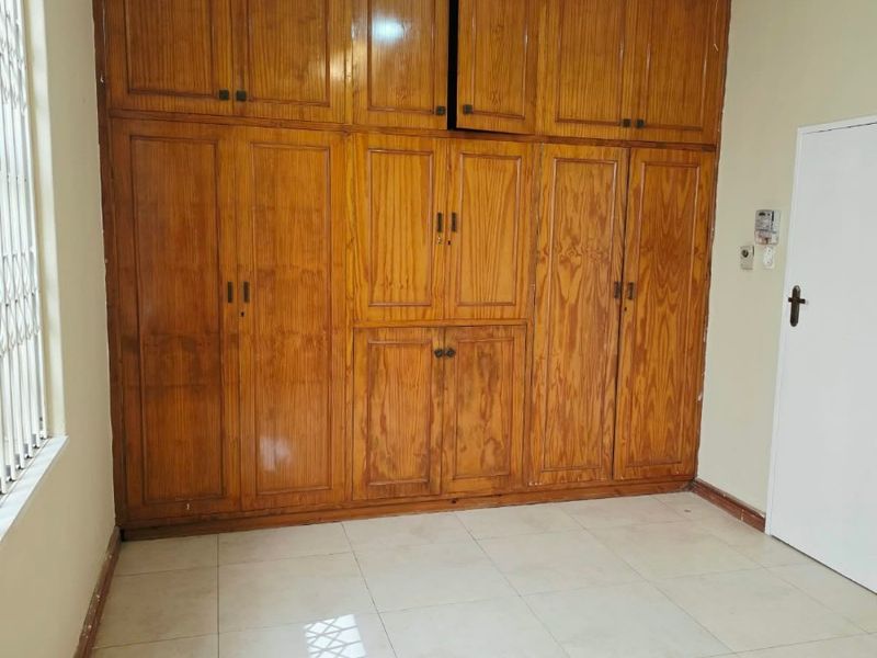 A bedroom featuring extensive floor-to-ceiling built-in wooden cupboards, neutral tiled flooring, and security bars on the window.