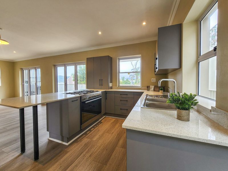 Modern open-plan kitchen featuring grey cabinetry, stone countertops, a gas hob, built-in oven, and double sink with views to the exterior.