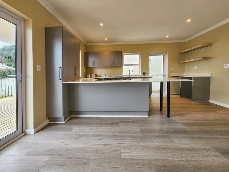 Modern open-plan kitchen featuring grey cabinetry, a central island with a gas hob, stone countertops, and light wood-look laminate flooring with direct patio access.