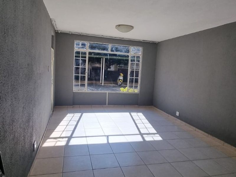 A spacious, empty living area featuring neutral tiled flooring, dark grey walls, and a large window providing natural light.