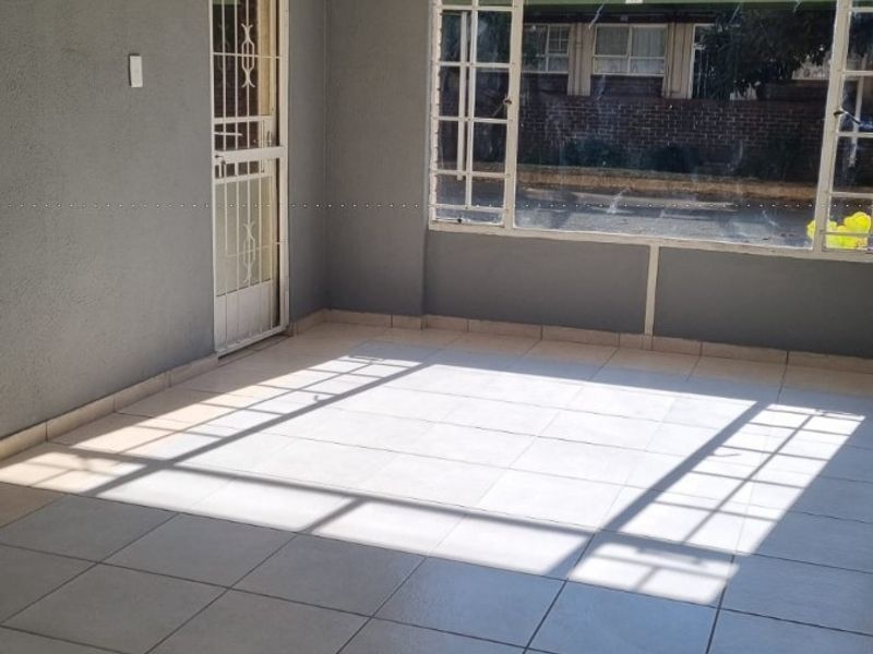 An empty, sunlit living area featuring neutral tiled flooring, a security gate, and a large window with a view of the exterior.