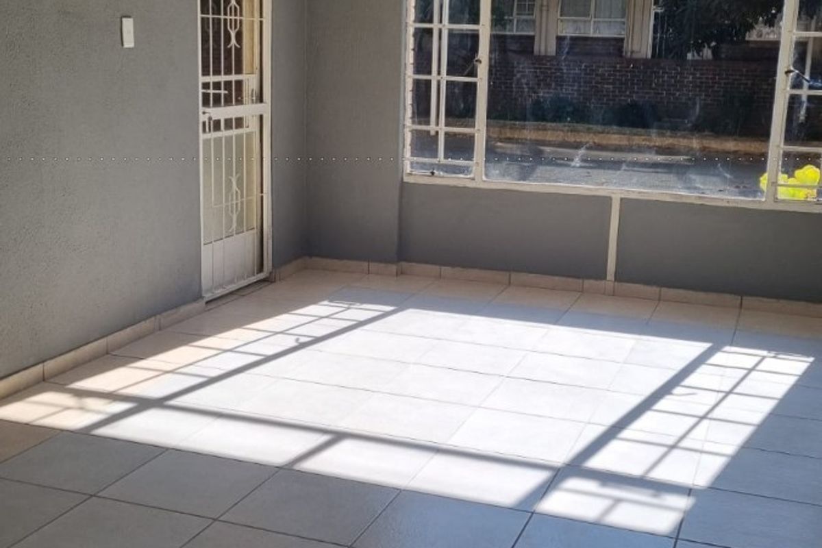 An empty, sunlit living area featuring neutral tiled flooring, a security gate, and a large window with a view of the exterior.