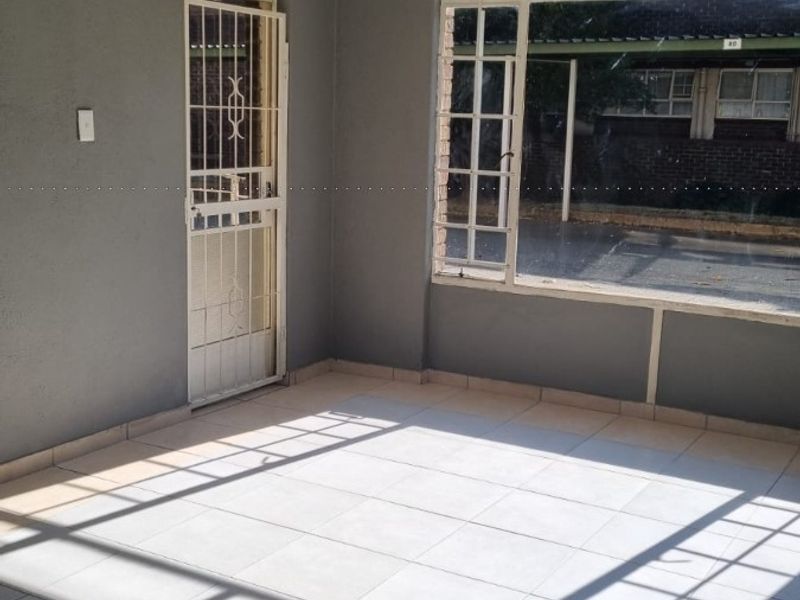 An empty, enclosed patio area featuring tiled flooring, a security gate, and large windows, offering a versatile space for relaxation or additional living area.