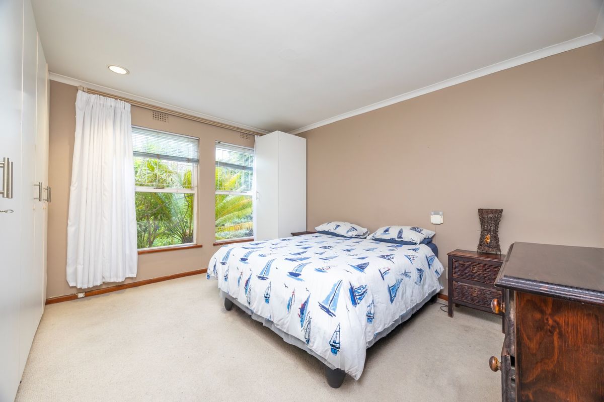 A well-lit bedroom featuring neutral walls, carpeted flooring, built-in cupboards, and large windows overlooking greenery.