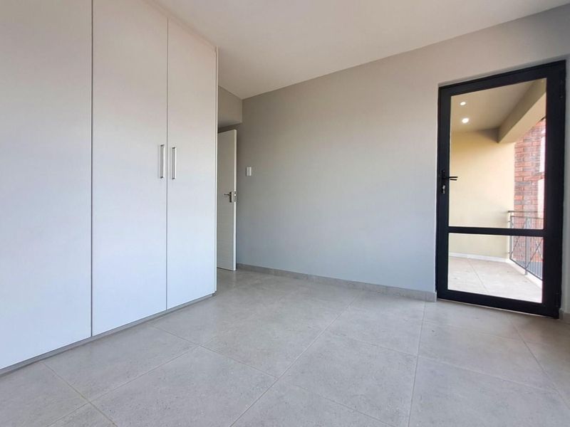 A bright, empty bedroom featuring neutral tiled flooring, built-in cupboards, and a glass-panelled door providing access to an outdoor balcony area.