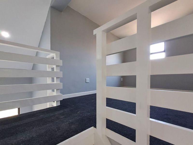 A bright, empty mezzanine hallway featuring clean grey walls, dark carpeting, and a white timber balustrade overlooking the lower level.