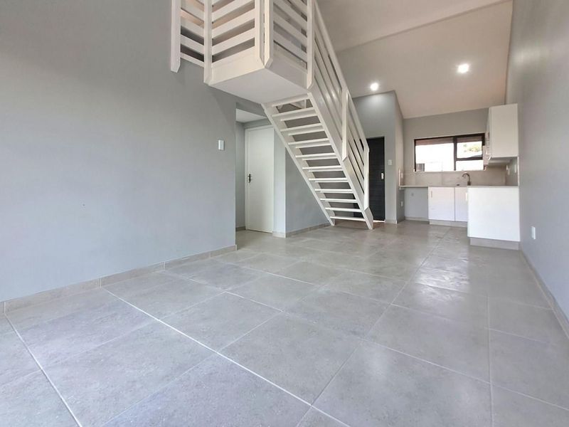Open-plan living area featuring tiled flooring, a modern staircase, and a compact kitchen with built-in cabinetry and sink.