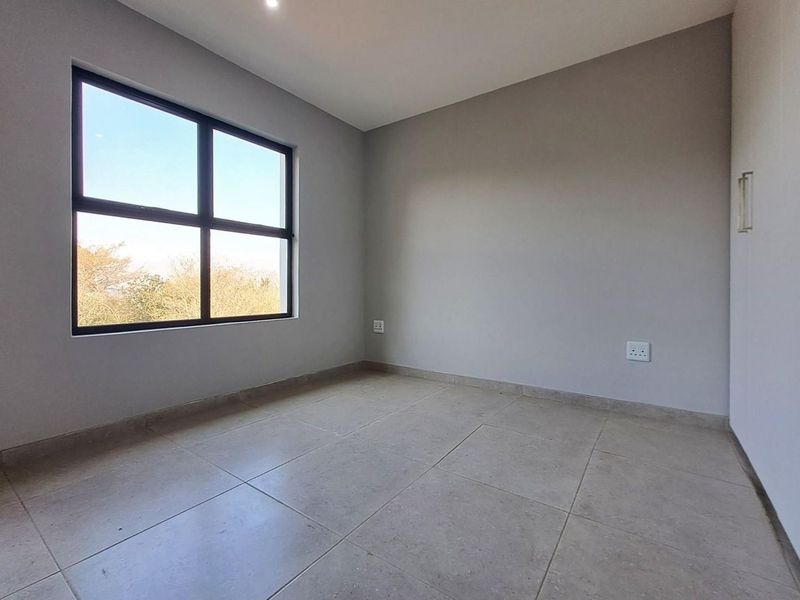 A bright, empty bedroom featuring neutral tiled flooring, a large black-framed window, and clean, modern finishes throughout.