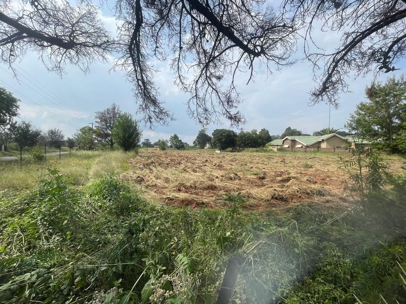 A view of a cleared, undeveloped plot of land on a farm property with surrounding vegetation and distant building structures.