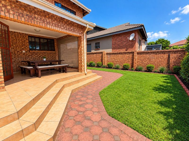 A well-maintained rear garden featuring a covered brick patio, tiled steps, paved walkway, and a neat lawn enclosed by a brick boundary wall.