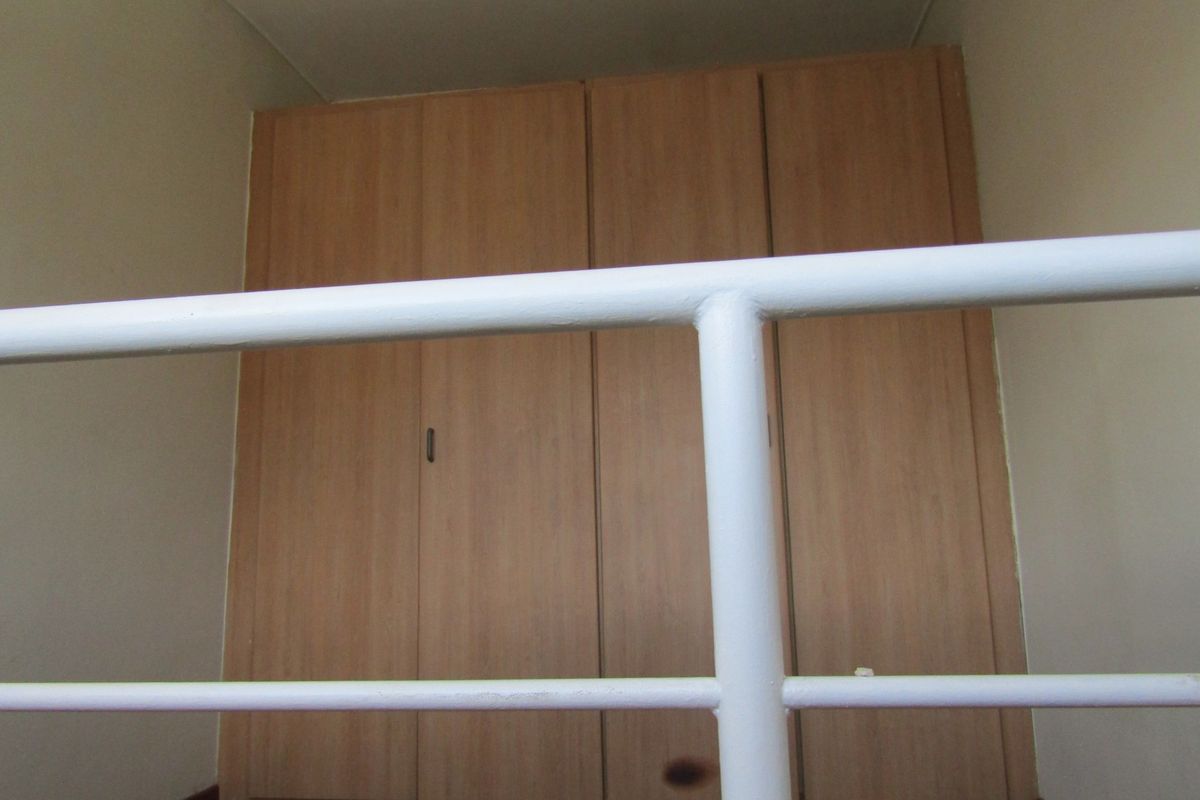 A bedroom interior featuring built-in wooden cupboards and a white metal balustrade, showing signs of wear.