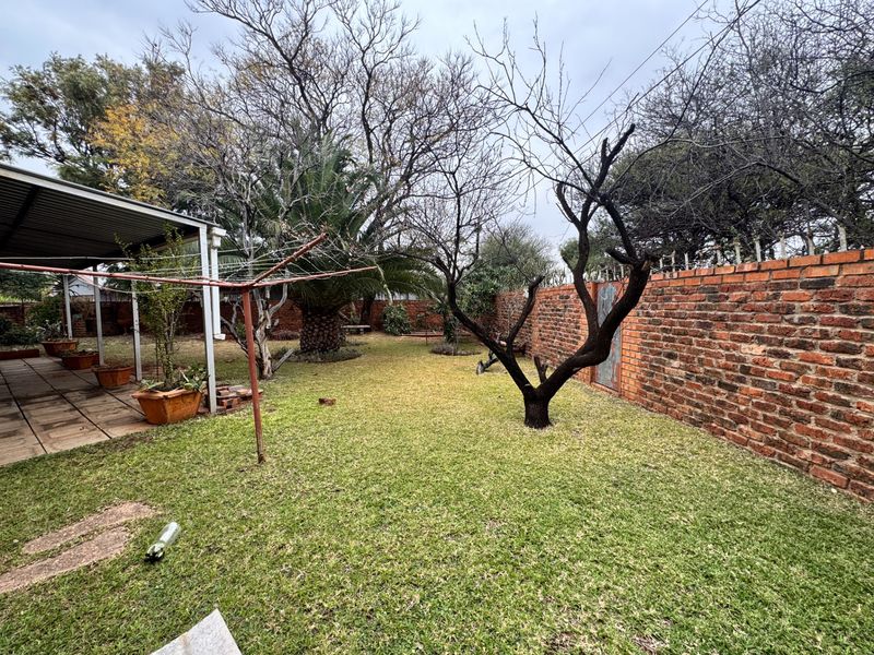 A private rear garden featuring a lawn, mature trees, a brick boundary wall, and an attached covered patio area.