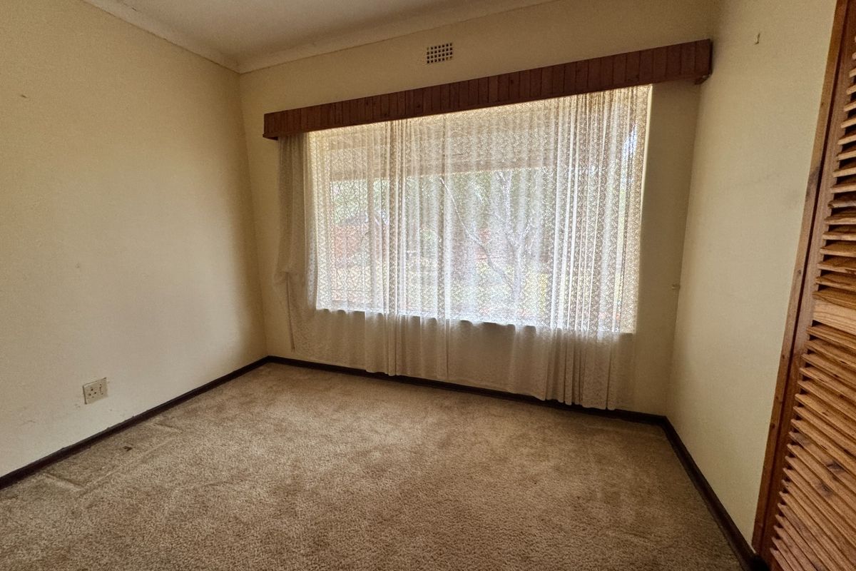 An empty bedroom featuring carpet flooring, a built-in wardrobe with louvered doors, and a large window with a decorative pelmet.