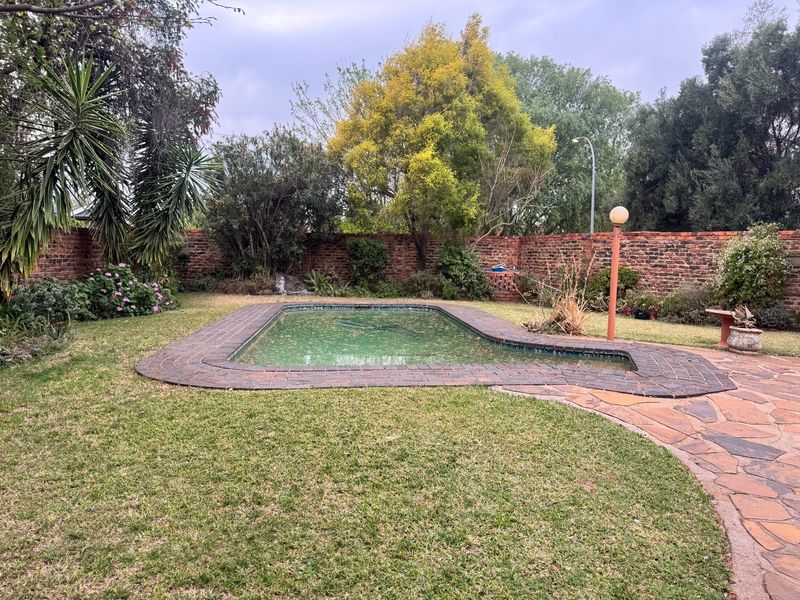 A private backyard featuring a swimming pool surrounded by a lawn, stone paving, and a brick boundary wall with mature garden landscaping.