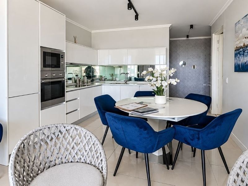 Modern open-plan kitchen featuring sleek white gloss cabinetry, integrated appliances, a mirrored splashback, and contemporary track lighting, seamlessly flowing into the dining area.