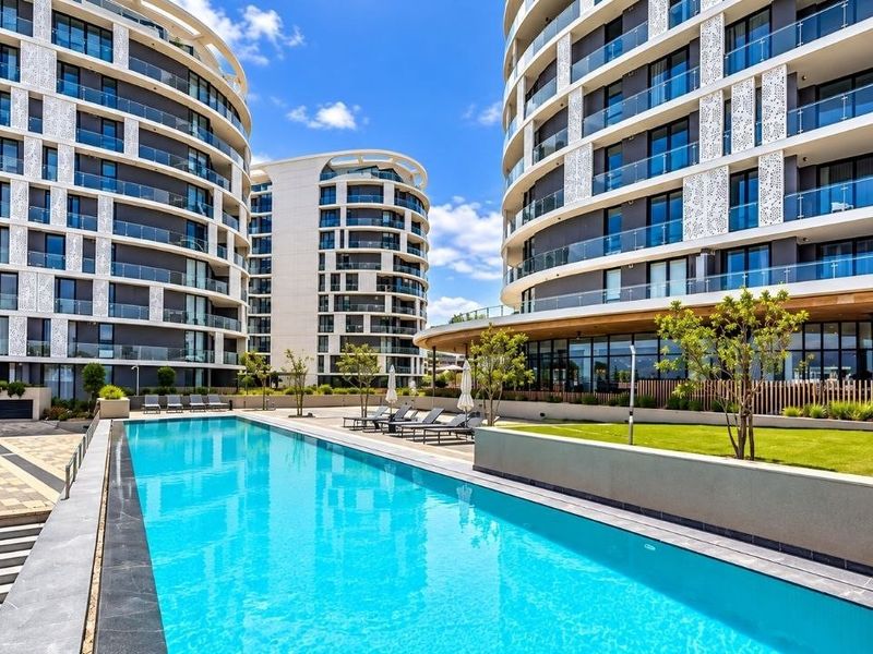A modern apartment complex featuring a pristine swimming pool, landscaped garden areas, and contemporary architectural facades with glass-fronted balconies.