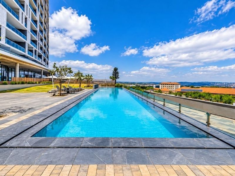 A modern infinity edge swimming pool featuring a glass balustrade, paved deck, and expansive views, set within a well-maintained apartment complex.
