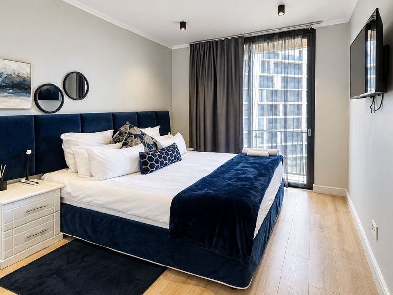 A well-maintained bedroom featuring light laminate flooring, recessed ceiling lighting, and direct access to a balcony through a sliding glass door.