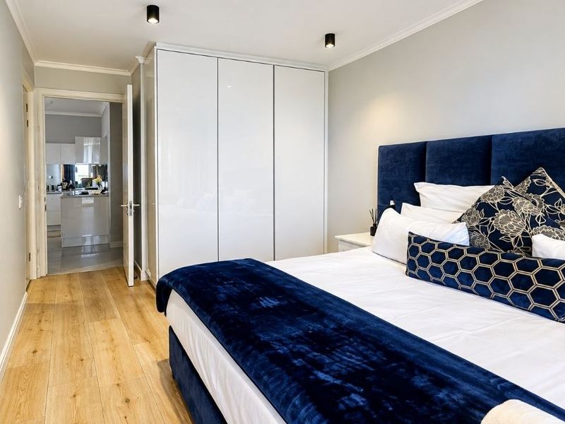 A modern, well-maintained bedroom featuring sleek built-in wardrobes, light laminate flooring, and recessed ceiling lighting, with an adjoining view into the kitchen area.