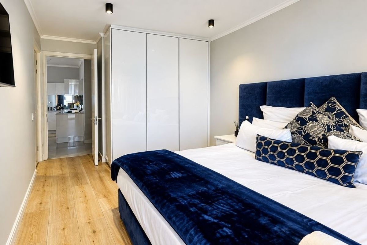 A modern, well-maintained bedroom featuring sleek built-in wardrobes, light laminate flooring, and recessed ceiling lighting, with an adjoining view into the kitchen area.