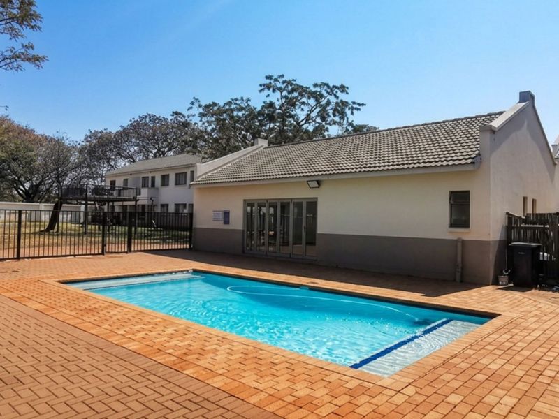 A well-maintained communal pool area featuring a paved deck, perimeter fencing, and access to a clubhouse building with tiled roofing.