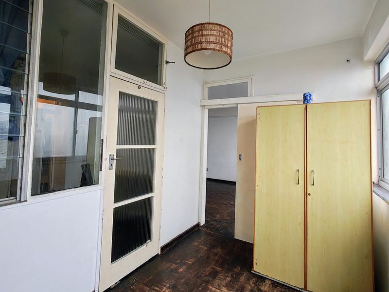 An enclosed balcony or sunroom featuring original parquet flooring, large windows, and a classic ribbed glass door leading to the interior.