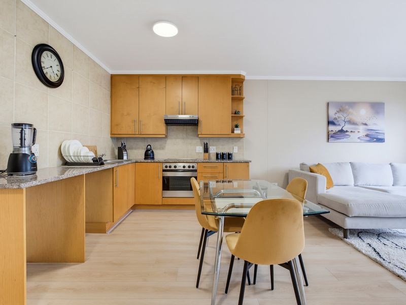 An open-plan living and kitchen area featuring light wood cabinetry, granite countertops, stainless steel appliances, and modern laminate flooring.
