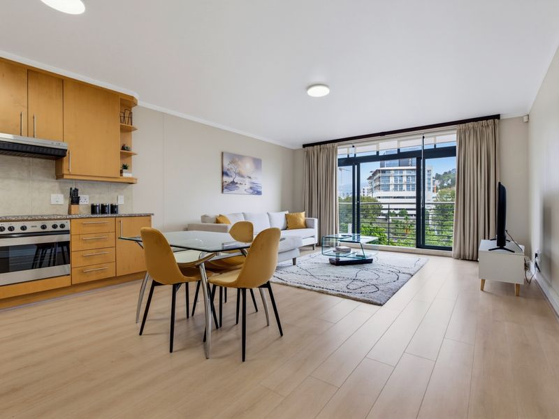 Open-plan living area featuring a functional kitchen with light wood cabinetry, laminate flooring, and large sliding doors leading to a balcony with city views.