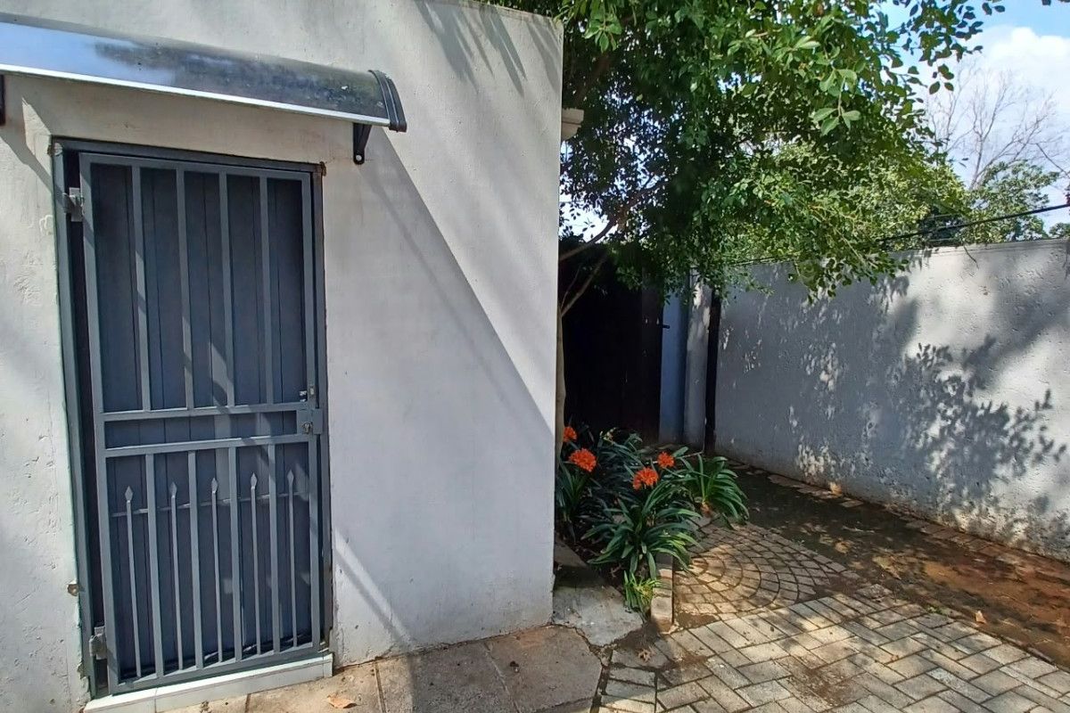 A rear exterior view of the cottage featuring a paved patio area, security gate, and a boundary wall with mature trees.