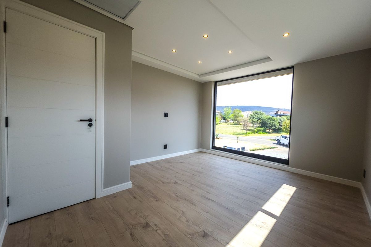 A bright, modern, and empty bedroom featuring laminate flooring, recessed ceiling lights, and a large window offering a pleasant exterior view.