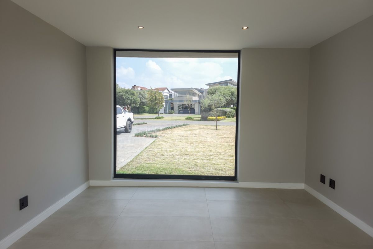 A bright, empty living room featuring large picture windows, neutral tiled flooring, and recessed downlighting, offering a clean and modern aesthetic.