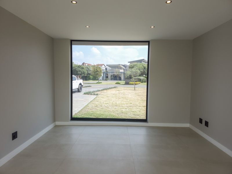 A bright, empty living room featuring large picture windows, neutral tiled flooring, and recessed downlighting, offering a clean and modern aesthetic.