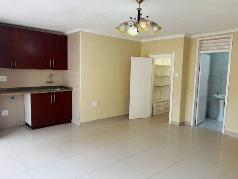An open-plan living area featuring tiled flooring, a compact kitchenette with built-in cabinetry, and access to a storage area and bathroom.