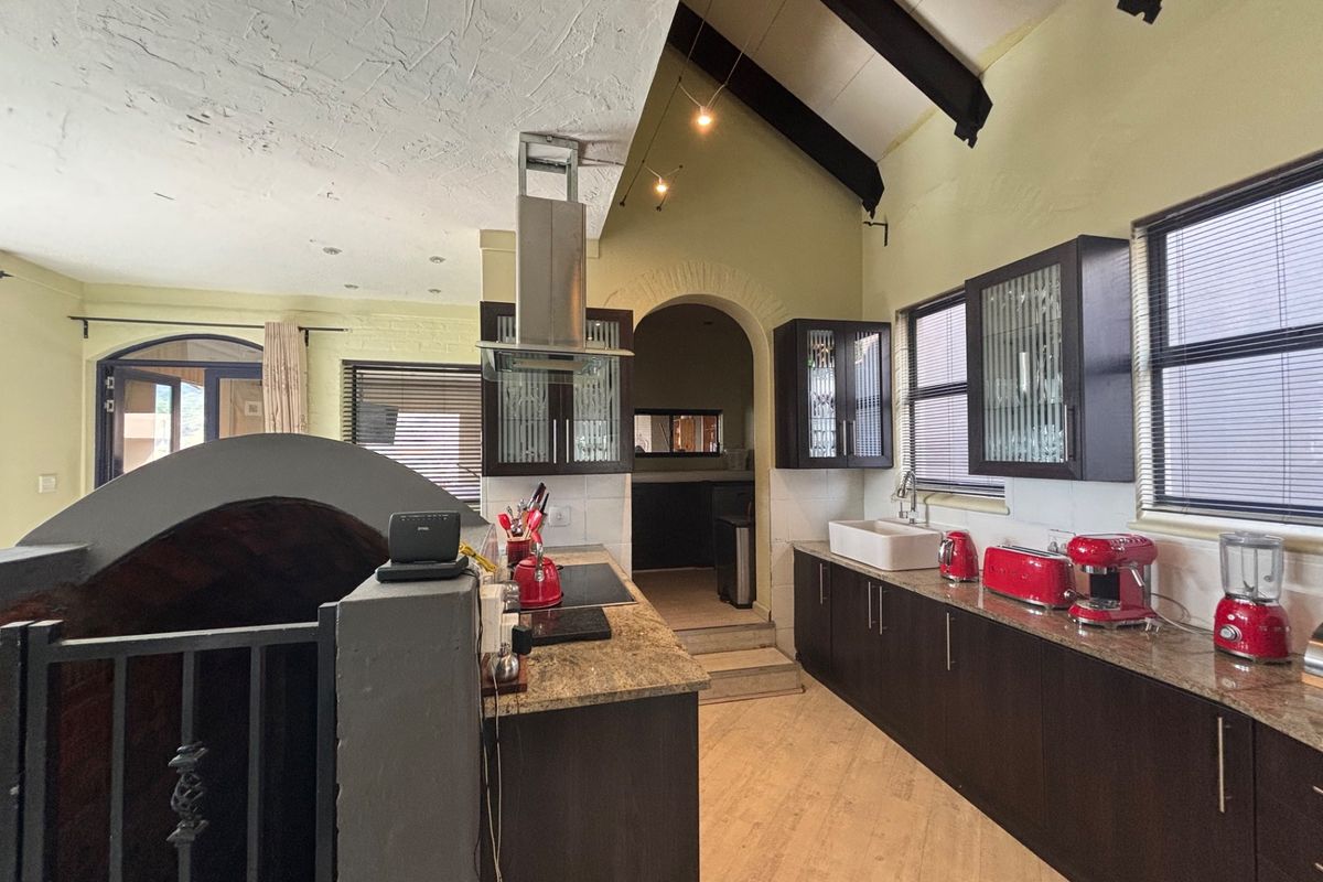A kitchen featuring dark wood cabinetry, granite countertops, a farmhouse sink, and a vaulted ceiling with exposed beams, leading to an arched doorway.