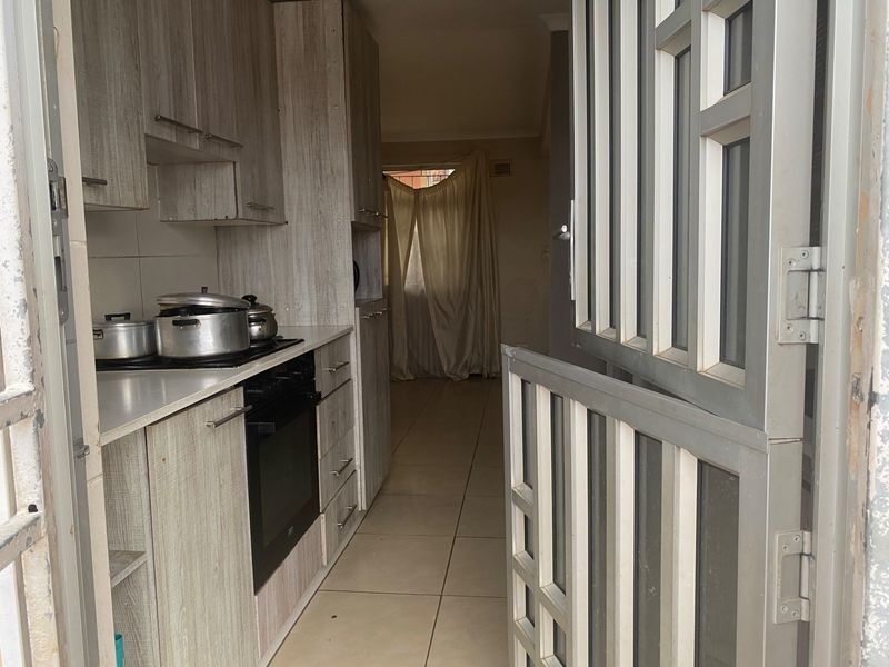 Compact kitchen featuring light wood-look cabinetry, built-in oven and hob, tiled flooring, and direct access through a security gate.