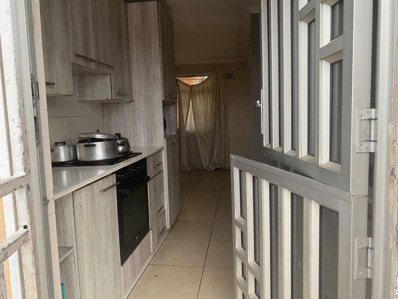A compact kitchen featuring light wood-grain cabinetry, built-in oven and hob, tiled flooring, and a secure metal entrance gate.
